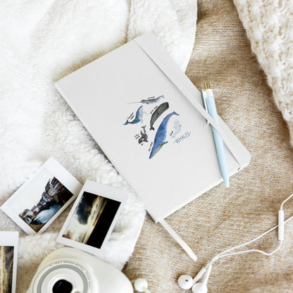 A white hardcover notebook with a blue whale illustration and the word 'WHALES' on the cover, styled in a cozy flat lay with a light blue pen, instant photos, earbuds, and a Polaroid camera on a beige textured blanket.